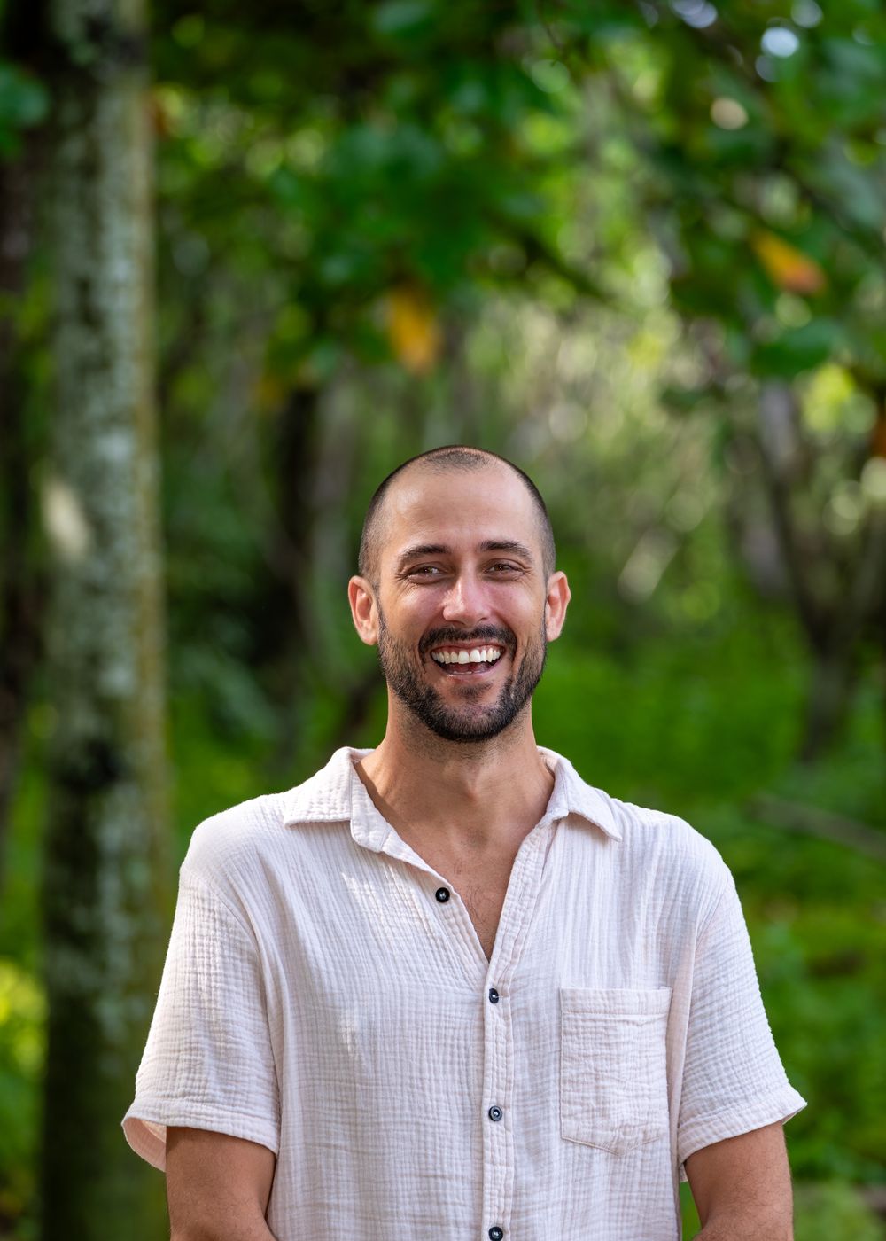 Kyle Bombard smiling, in a cream linen shirt, with green forest behind him